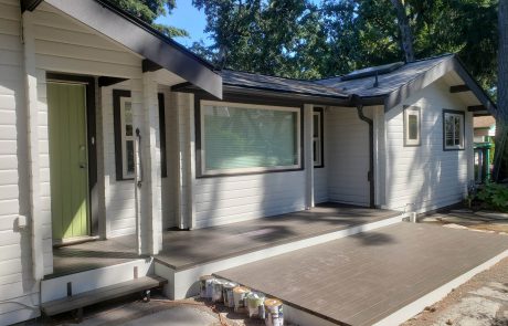 a white house with black roof and window frames surrounded by trees and has a stained front deck